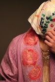 A person is partially covered with a floral patterned scarf over their head and wearing a draped garment with a rose design. The garment is in shades of pink, and the scarf is white with multicolored floral prints. Jewelry with multiple chains is visible on the person's neck.