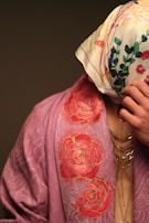 A person is partially covered with a floral patterned scarf over their head and wearing a draped garment with a rose design. The garment is in shades of pink, and the scarf is white with multicolored floral prints. Jewelry with multiple chains is visible on the person's neck.