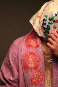 A person is partially covered with a floral patterned scarf over their head and wearing a draped garment with a rose design. The garment is in shades of pink, and the scarf is white with multicolored floral prints. Jewelry with multiple chains is visible on the person's neck.