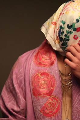 A person is partially covered with a floral patterned scarf over their head and wearing a draped garment with a rose design. The garment is in shades of pink, and the scarf is white with multicolored floral prints. Jewelry with multiple chains is visible on the person's neck.