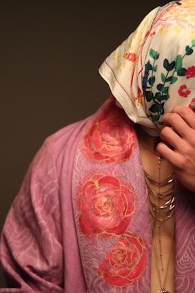 A person is partially covered with a floral patterned scarf over their head and wearing a draped garment with a rose design. The garment is in shades of pink, and the scarf is white with multicolored floral prints. Jewelry with multiple chains is visible on the person's neck.