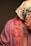 A person is partially covered with a floral patterned scarf over their head and wearing a draped garment with a rose design. The garment is in shades of pink, and the scarf is white with multicolored floral prints. Jewelry with multiple chains is visible on the person's neck.