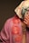 A person is partially covered with a floral patterned scarf over their head and wearing a draped garment with a rose design. The garment is in shades of pink, and the scarf is white with multicolored floral prints. Jewelry with multiple chains is visible on the person's neck.