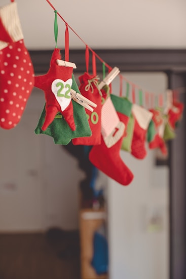A string of Christmas-themed decorations hangs across the room, featuring red and green felt stockings and pouches, each adorned with numbers and simple patterns. Clothespins are used to attach the decorations to the ribbon, which is strung along a wall or through a doorway. The scene evokes a festive and cozy atmosphere.