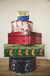 A stack of decorated holiday-themed tins, featuring various patterns. The top tin is small, blue with a gold lid, and has a gingerbread figure. The second tin is round with red and white foliage designs. Below it is a rectangular tin covered in a red pattern. The fourth tin is rectangular, predominantly green, with images of Christmas stockings and ornaments. The bottom tin is circular with a black background and silver snowflake designs.