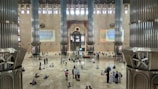A vast and spacious interior of a mosque with large, ornate pillars. The floor is made of polished tiles, and there are many people scattered around, some sitting, others standing. The wall features intricate designs and calligraphy. Light enters through high windows, creating a serene atmosphere.