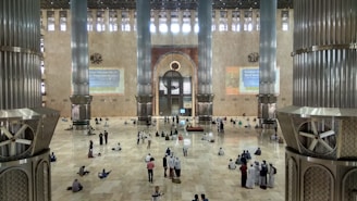 A vast and spacious interior of a mosque with large, ornate pillars. The floor is made of polished tiles, and there are many people scattered around, some sitting, others standing. The wall features intricate designs and calligraphy. Light enters through high windows, creating a serene atmosphere.