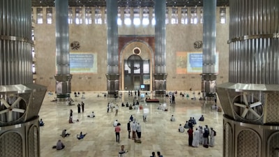 A vast and spacious interior of a mosque with large, ornate pillars. The floor is made of polished tiles, and there are many people scattered around, some sitting, others standing. The wall features intricate designs and calligraphy. Light enters through high windows, creating a serene atmosphere.