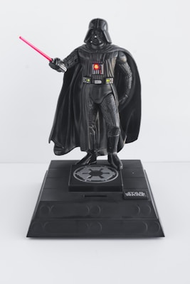 A detailed figure of a character in a dark, armored costume holding a red lightsaber. The character is positioned on a stylized black pedestal with a sci-fi emblem and a recognizable logo on the front.