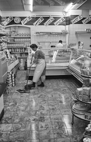 A black and white scene depicting a grocery store interior with a tiled floor. A person is mopping the floor near a refrigerated display case, and another person stands behind a counter. Various products line the shelves, and sales signs with prices hang above.