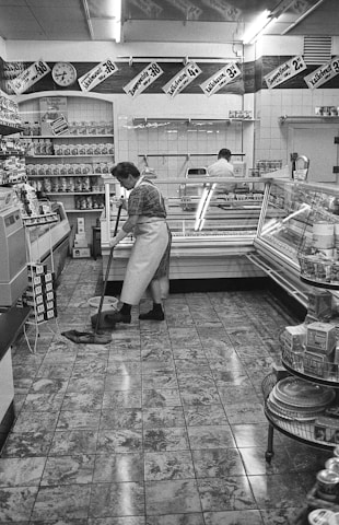 A black and white scene depicting a grocery store interior with a tiled floor. A person is mopping the floor near a refrigerated display case, and another person stands behind a counter. Various products line the shelves, and sales signs with prices hang above.
