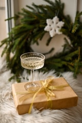 A crystal wine glass is placed on top of a brown gift box wrapped with a gold ribbon. Behind the gift box is a green Christmas wreath adorned with a white snowflake decoration. The setting is on a fluffy white surface, suggesting a cozy festive ambiance.