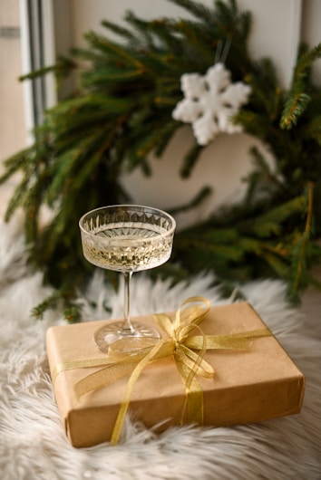 A crystal wine glass is placed on top of a brown gift box wrapped with a gold ribbon. Behind the gift box is a green Christmas wreath adorned with a white snowflake decoration. The setting is on a fluffy white surface, suggesting a cozy festive ambiance.