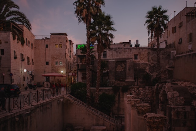 An ancient stone building is situated among palm trees in an urban setting. The architecture features a series of arches and old stonework, with a rustic, weathered appearance. There are street lamps and minimal vehicle presence suggesting a calm atmosphere at dusk. A warm, dim light from the windows adds to the serene ambiance, while neon signs in Hebrew add a touch of modernity to the historical surroundings.