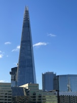A tall, pointed skyscraper dominates the skyline against a clear blue sky. The building is modern, with glass panels reflecting light. Surrounding structures include other modern buildings, some with curved designs and one with the name 'London Bridge Hospital' visible.