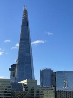 A tall, pointed skyscraper dominates the skyline against a clear blue sky. The building is modern, with glass panels reflecting light. Surrounding structures include other modern buildings, some with curved designs and one with the name 'London Bridge Hospital' visible.