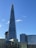 A tall, pointed skyscraper dominates the skyline against a clear blue sky. The building is modern, with glass panels reflecting light. Surrounding structures include other modern buildings, some with curved designs and one with the name 'London Bridge Hospital' visible.