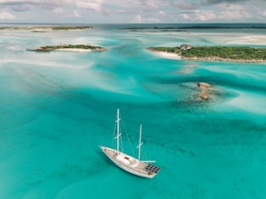 A tranquil scene with a turquoise sea surrounding a small, lush island. A solitary sailboat is anchored in the clear waters, and a wooden dock extends from the island, which features a small building nestled among greenery. The sky is partly cloudy, adding depth to the peaceful maritime landscape.