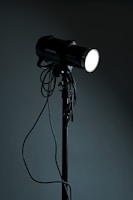 A studio spotlight on a tripod stand with various cables hanging from it. The light is turned on, emitting a bright glow in contrast with the dark background.