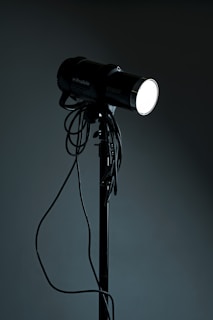 A studio spotlight on a tripod stand with various cables hanging from it. The light is turned on, emitting a bright glow in contrast with the dark background.