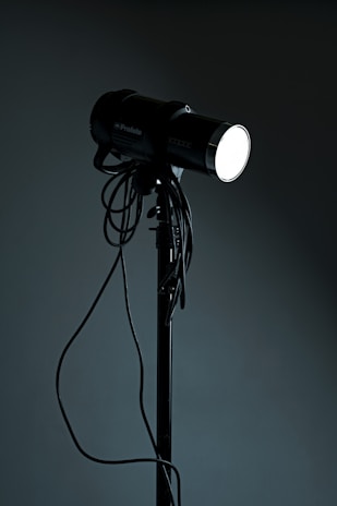 A studio spotlight on a tripod stand with various cables hanging from it. The light is turned on, emitting a bright glow in contrast with the dark background.