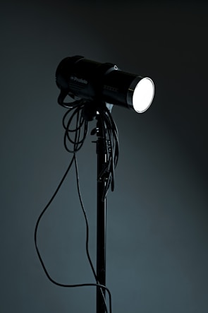 A studio spotlight on a tripod stand with various cables hanging from it. The light is turned on, emitting a bright glow in contrast with the dark background.
