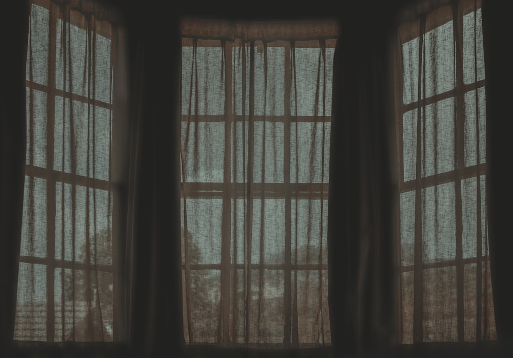 a dark room with two open windows with sheer curtains