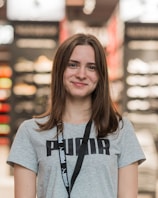 A young woman with long brown hair is smiling while wearing a gray Puma t-shirt and a lanyard. She is standing indoors, and the background appears to be a store, with blurred shelves and lights.