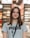 A young woman with long brown hair is smiling while wearing a gray Puma t-shirt and a lanyard. She is standing indoors, and the background appears to be a store, with blurred shelves and lights.