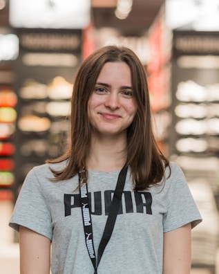 A young woman with long brown hair is smiling while wearing a gray Puma t-shirt and a lanyard. She is standing indoors, and the background appears to be a store, with blurred shelves and lights.