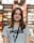 A young woman with long brown hair is smiling while wearing a gray Puma t-shirt and a lanyard. She is standing indoors, and the background appears to be a store, with blurred shelves and lights.