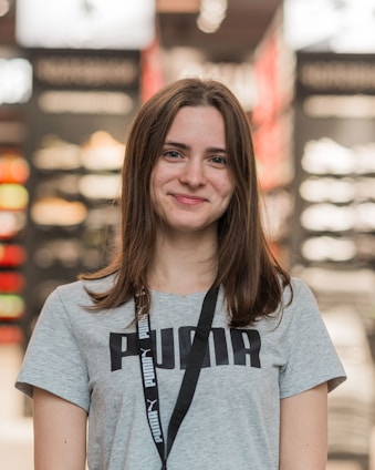 A young woman with long brown hair is smiling while wearing a gray Puma t-shirt and a lanyard. She is standing indoors, and the background appears to be a store, with blurred shelves and lights.