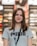 A young woman with long brown hair is smiling while wearing a gray Puma t-shirt and a lanyard. She is standing indoors, and the background appears to be a store, with blurred shelves and lights.