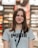 A young woman with long brown hair is smiling while wearing a gray Puma t-shirt and a lanyard. She is standing indoors, and the background appears to be a store, with blurred shelves and lights.