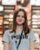A young woman with long brown hair is smiling while wearing a gray Puma t-shirt and a lanyard. She is standing indoors, and the background appears to be a store, with blurred shelves and lights.