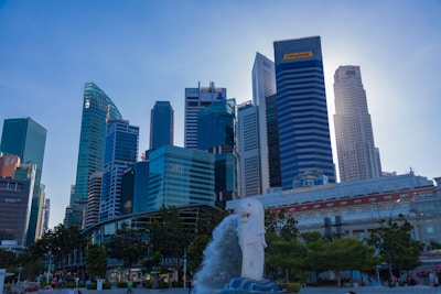 A cityscape featuring a skyline with modern skyscrapers of various architectural styles, dominated by glass and steel facades. In the foreground, there is a large white statue of a mythical creature that appears to be a Merlion, which is spouting water into the air. Plush greenery and trees are visible in the vicinity, creating a blend of urban and natural elements.