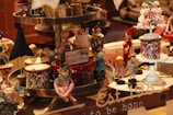 A festive display of Christmas-themed decorations featuring wooden toy figures in vibrant costumes, drums with intricate geometric patterns, a small white Christmas tree adorned with green baubles, and a sign reading 'Merry Christmas'. There's a cozy, nostalgic atmosphere with a mix of colors and textures.