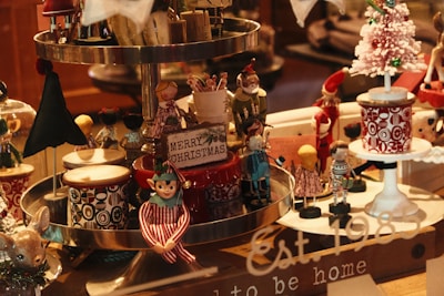 A festive display of Christmas-themed decorations featuring wooden toy figures in vibrant costumes, drums with intricate geometric patterns, a small white Christmas tree adorned with green baubles, and a sign reading 'Merry Christmas'. There's a cozy, nostalgic atmosphere with a mix of colors and textures.