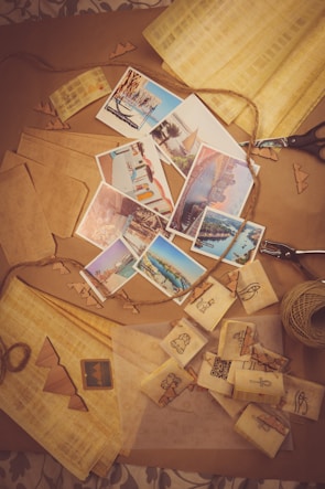 A collection of postcards depicting various tourist destinations is spread across a table. Surrounding the postcards are items associated with crafts such as twine, paper cutouts, and scissors. The scene suggests an activity of creating or packaging postcards with a focus on travel and keepsakes.