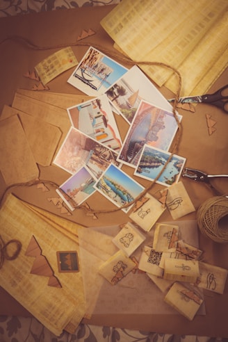 A collection of postcards depicting various tourist destinations is spread across a table. Surrounding the postcards are items associated with crafts such as twine, paper cutouts, and scissors. The scene suggests an activity of creating or packaging postcards with a focus on travel and keepsakes.