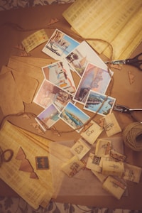 A collection of postcards depicting various tourist destinations is spread across a table. Surrounding the postcards are items associated with crafts such as twine, paper cutouts, and scissors. The scene suggests an activity of creating or packaging postcards with a focus on travel and keepsakes.