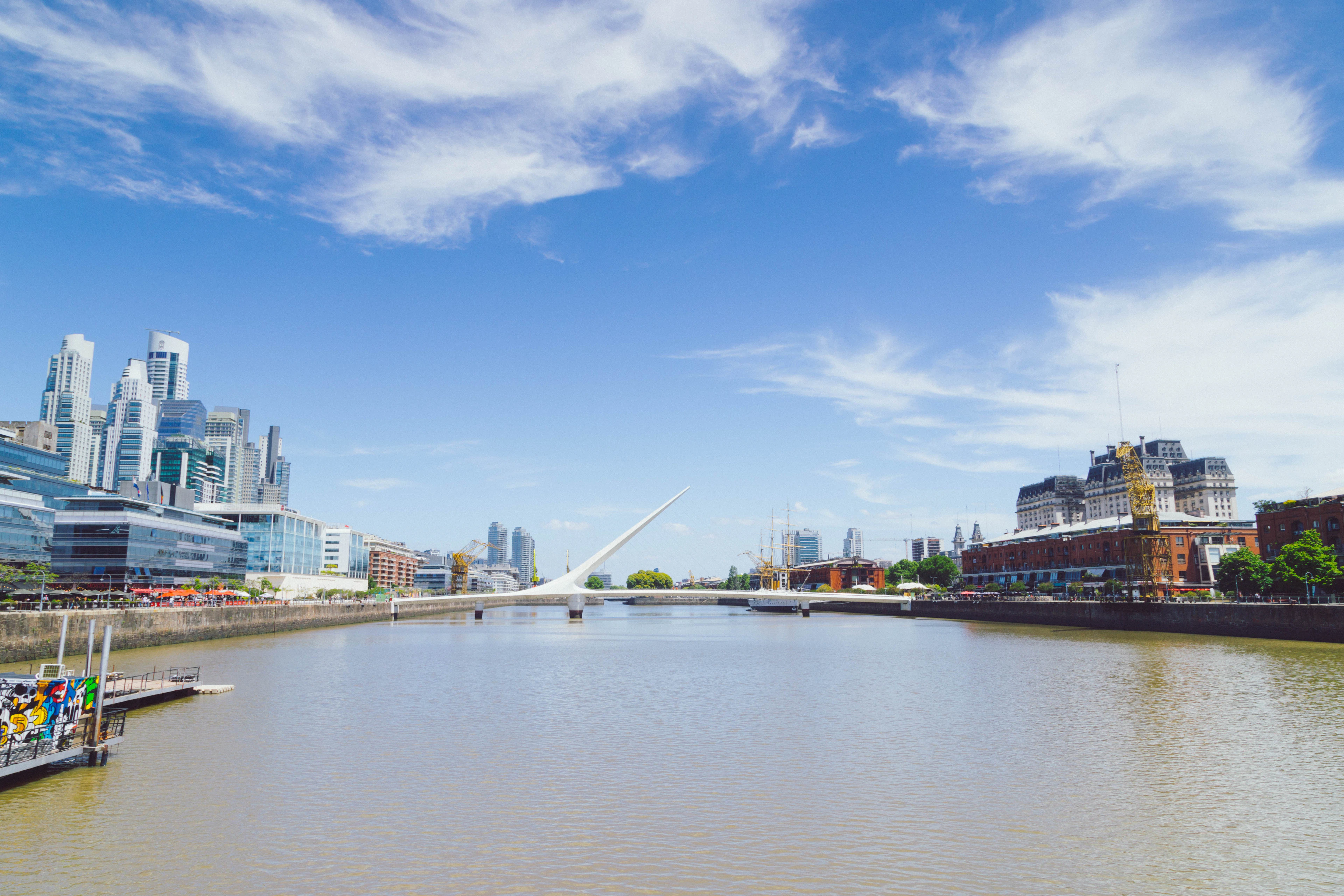 The white Puente de la Mujer footbridge spanning a dock in Buenos Aires' Puerto Madero, glass office towers along one quay and restored red-brick warehouses along the other under a bright blue sky.