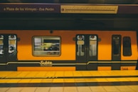 A bright orange subway train is stationed at an underground platform, with passengers visible through the windows. The train is branded 'Subte' indicating it's part of the Buenos Aires Metro. Overhead, the signboard displays the name 'Plaza de los Virreyes - Eva Perón' with a route diagram. The platform is marked with yellow and black caution lines.