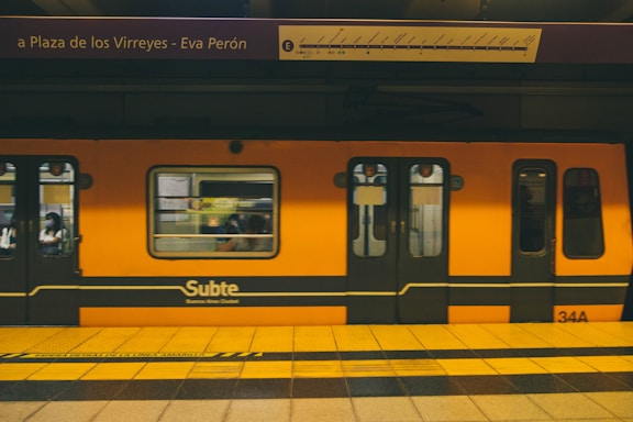 A bright orange subway train is stationed at an underground platform, with passengers visible through the windows. The train is branded 'Subte' indicating it's part of the Buenos Aires Metro. Overhead, the signboard displays the name 'Plaza de los Virreyes - Eva Perón' with a route diagram. The platform is marked with yellow and black caution lines.
