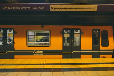 A bright orange subway train is stationed at an underground platform, with passengers visible through the windows. The train is branded 'Subte' indicating it's part of the Buenos Aires Metro. Overhead, the signboard displays the name 'Plaza de los Virreyes - Eva Perón' with a route diagram. The platform is marked with yellow and black caution lines.