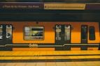 A bright orange subway train is stationed at an underground platform, with passengers visible through the windows. The train is branded 'Subte' indicating it's part of the Buenos Aires Metro. Overhead, the signboard displays the name 'Plaza de los Virreyes - Eva Perón' with a route diagram. The platform is marked with yellow and black caution lines.