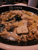 A bowl of steaming tofu and vegetable soup, featuring pieces of tofu, greens, and a creamy broth. The dish appears to be garnished with sesame seeds and sits in a bowl labeled 'Jinya'. The soup is hearty and rich, with a mix of textures from the vegetables and tofu.