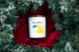 A candle with a white label and a brown lid is placed on a bright red fabric, surrounded by green pine branches with white snow-like accents. The candle label has modern typography and minimalistic design elements.