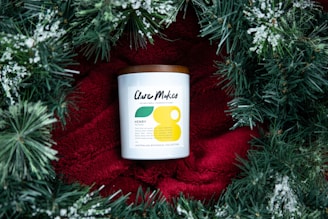 A candle with a white label and a brown lid is placed on a bright red fabric, surrounded by green pine branches with white snow-like accents. The candle label has modern typography and minimalistic design elements.