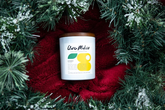 A candle with a white label and a brown lid is placed on a bright red fabric, surrounded by green pine branches with white snow-like accents. The candle label has modern typography and minimalistic design elements.
